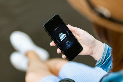 A person holding a black smartphone showing a digital screen, with their hand, legs, and a brown hat visible in a softly blurred real-world setting.